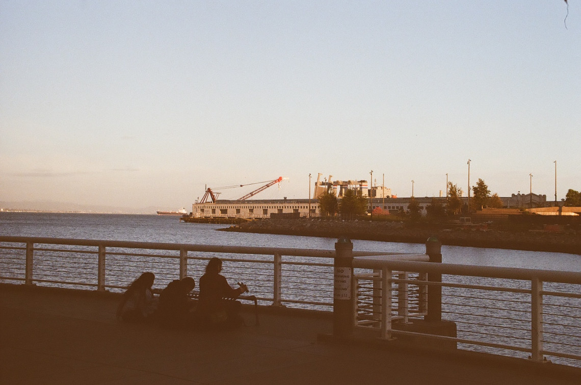 A group of 3 people sitting at a bench next to the oceanside. A large ship is visible.